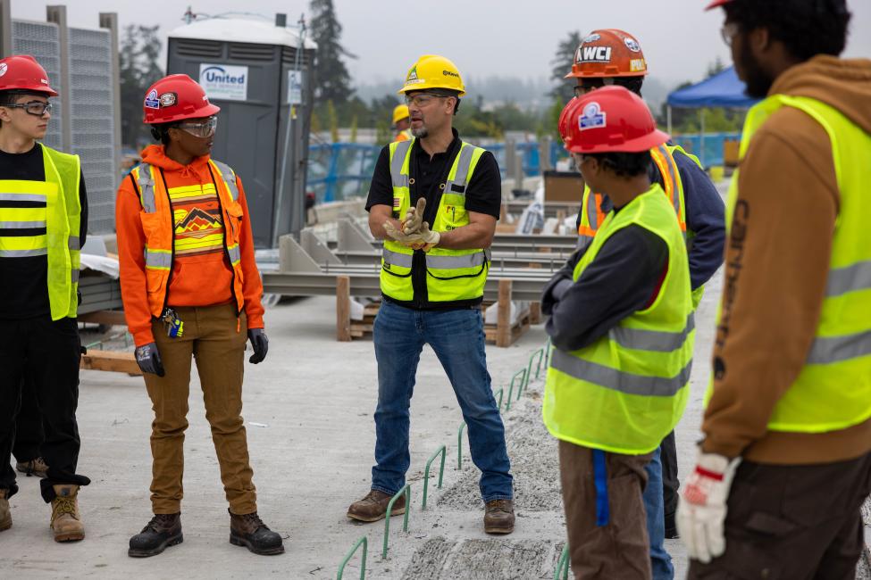 A construction worker explains a concept to young people of color touring the site. 
