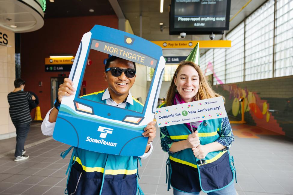 Two Sound Transit employees smile while holding up signs celebrating the one year anniversary of Roosevelt Station.