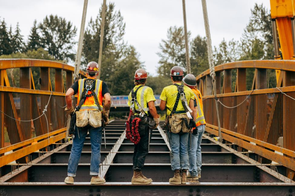 Four workers stand on the bridge, with their backs to the camera. 