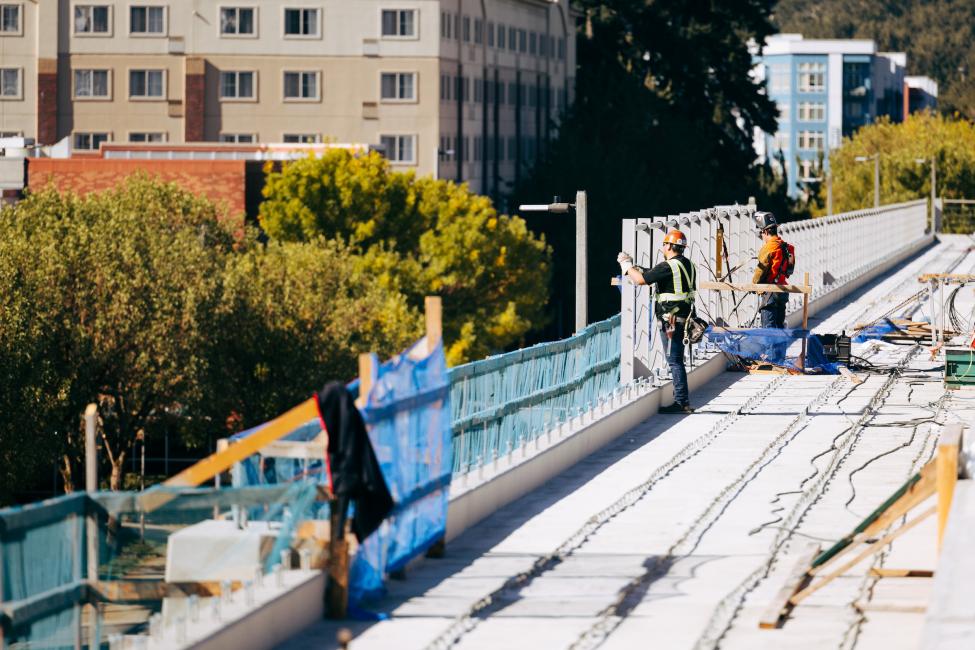 Two workers on an elevated light rail guideway in downtown Redmond, with green trees in the background. 