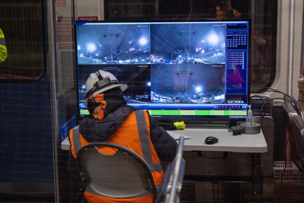 A person wearing a hard hat and orange vest looks at four computer screens.