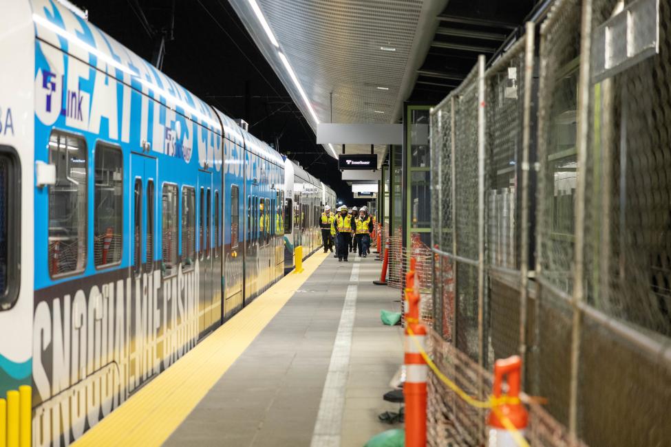 A group of people wearing hard hats and yellow jackets walk next to a Link train.