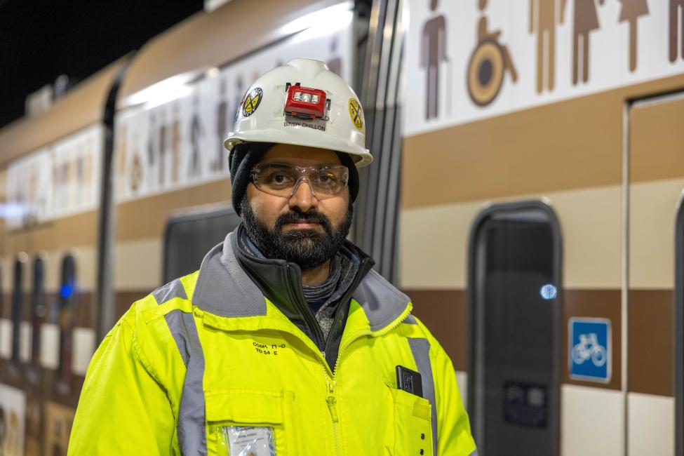 A crew member looks at the camera, wearing a yellow jacket and hard hat with a Link train in the background.