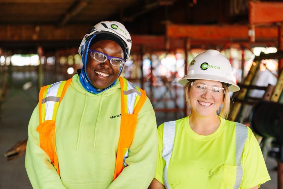Two women wearing bright colors and hard hats smile. 
