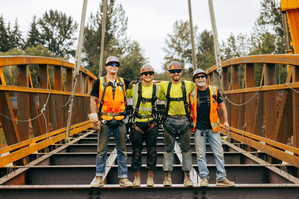 A group of four construction workers put their hands around each others' backs and smile at the camera while standing on a partially completed bridge. 