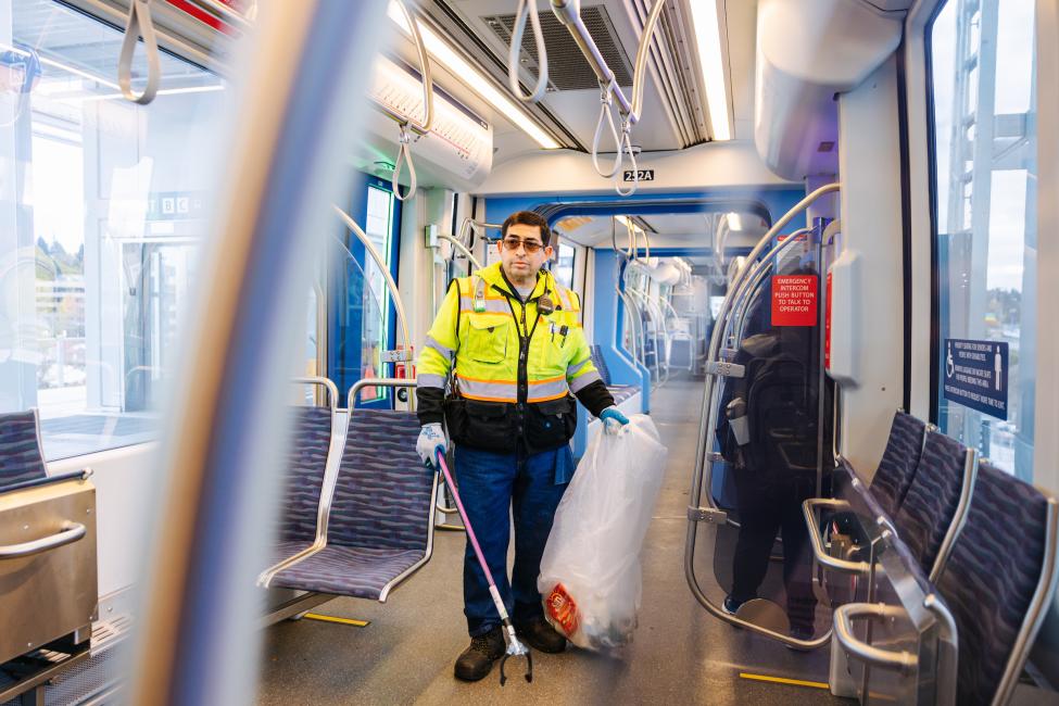 A cleaner wearing a bright yellow jacket and holding a garbage bag walks through a Link train. 