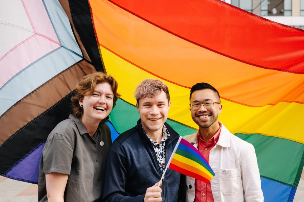 Three people pose in front of the Progress Pride flag. 