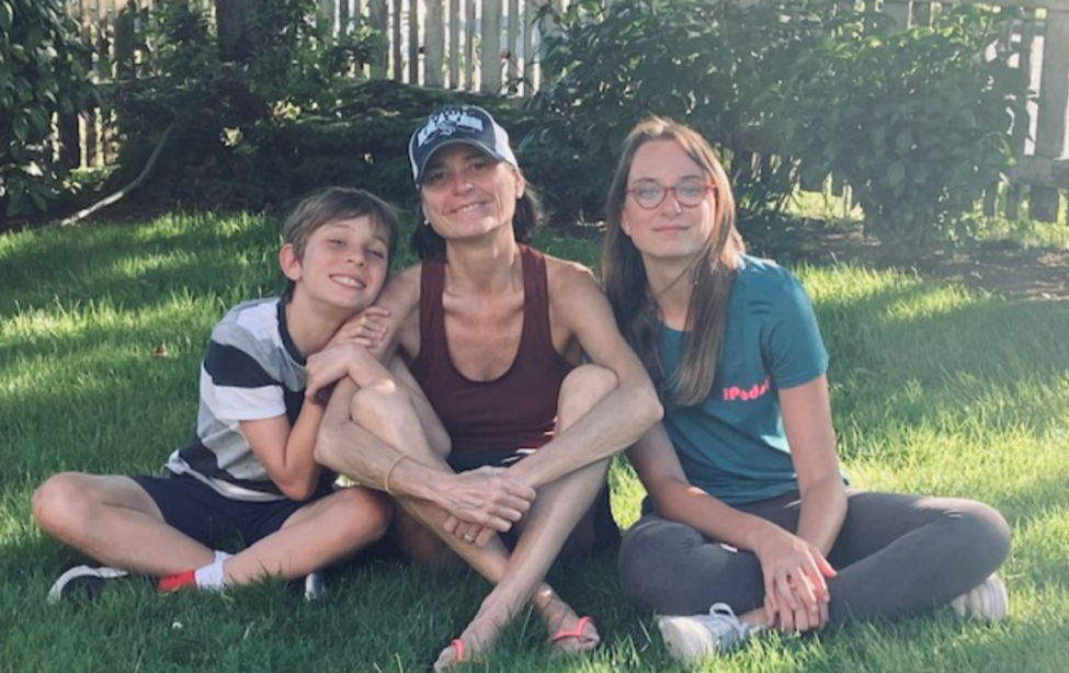 Clara O'Brien and her kids smile while sitting cross-legged on a grass lawn.