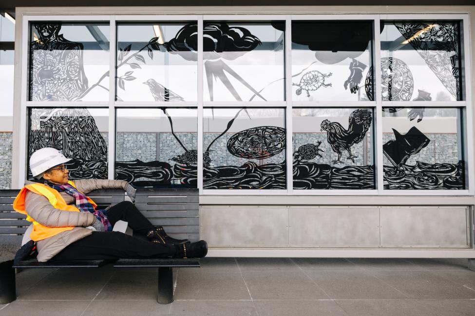 Barbara Earl Thomas sits on a bench at Judkins Park Station, in front of the windscreens she designed. She wears an orange vest and hard hat, as the station is still under construction. 