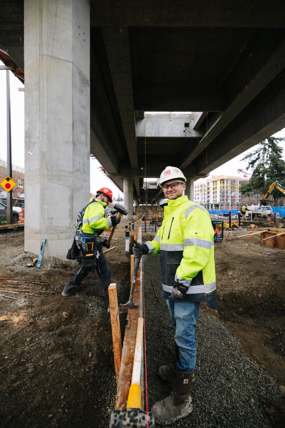 Two workers in hard hats and yellow jackets smile. 