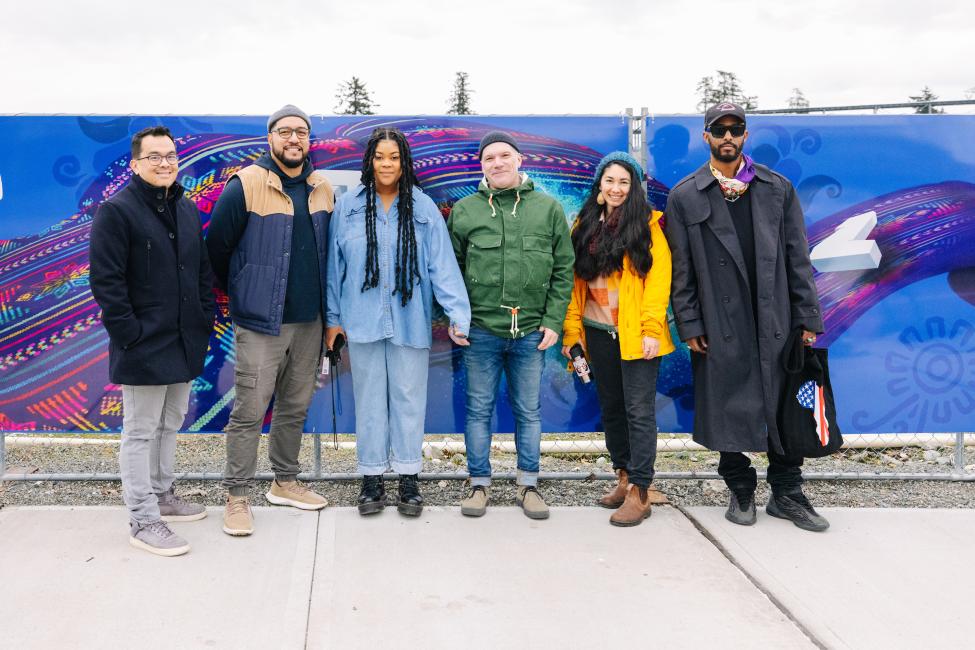 Six artists smile while standing in front of an art piece attached to a construction fence