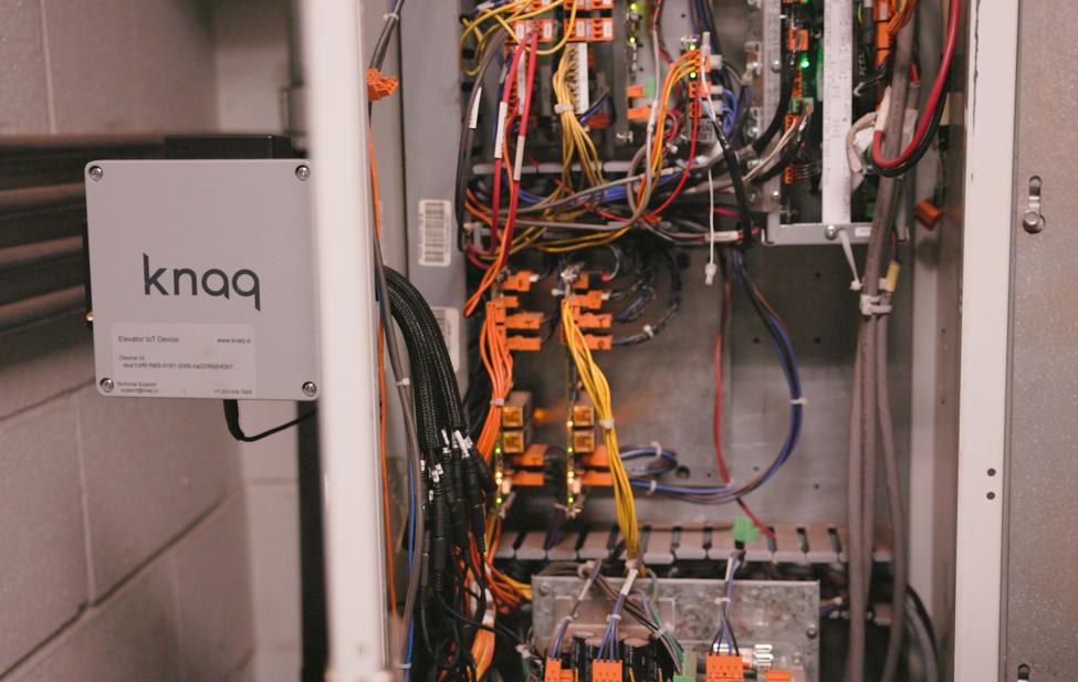 A box with text 'Knaq' is next to an elevator control unit. It is a piece of equipment that monitors and collects data on the elevator performance