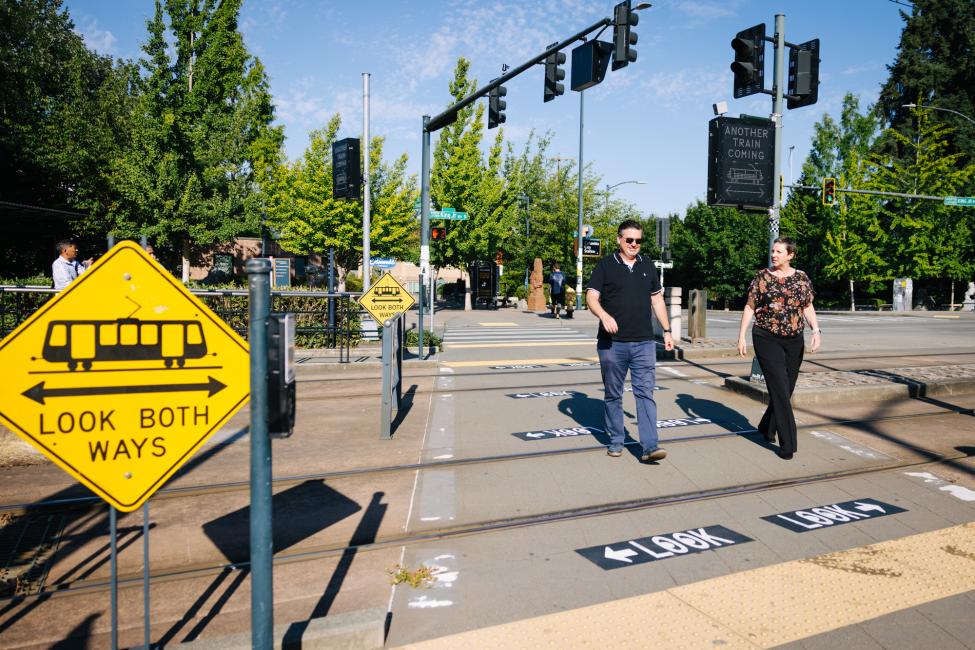 Two people cross the street over light rail tracks in the Rainier Valley, with new safety signage on the pavement reading 'Look'