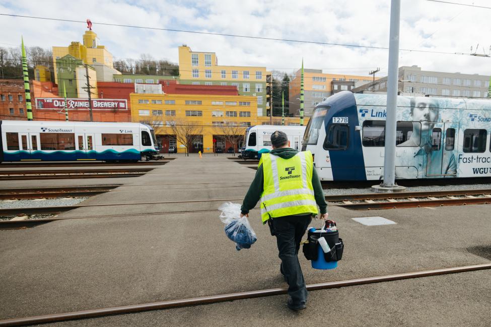 A person wearing a yellow vest and holding cleaning supplies walks through the Link train yard in South Seattle.