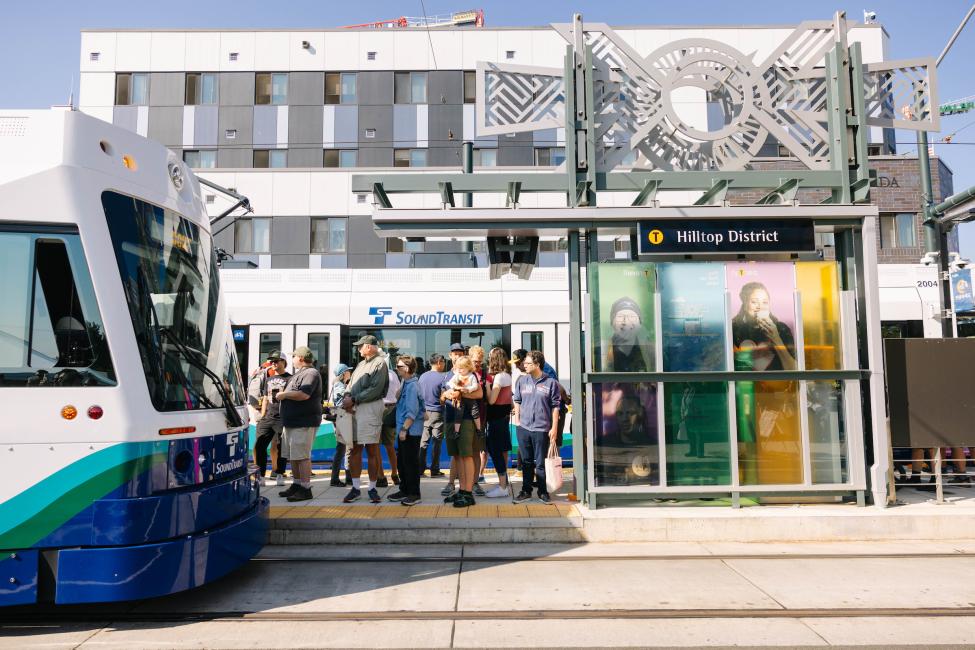 People wait on the platform as a T Line vehicle pulls in to Hilltop Station on a sunny day