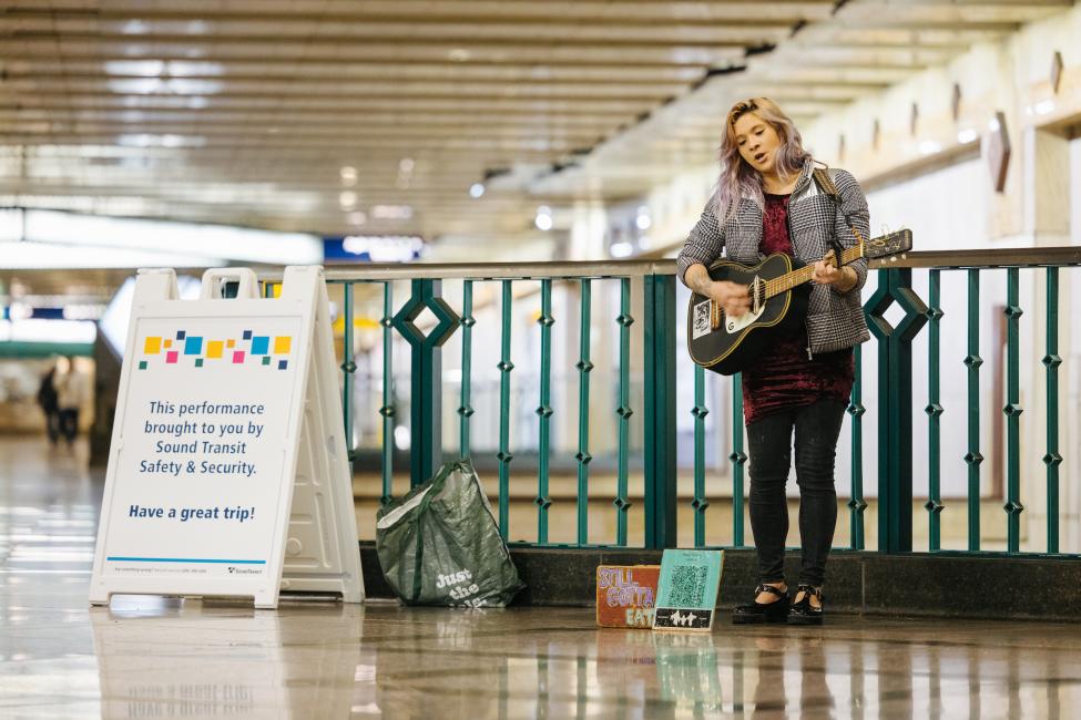 Ruby stands with her guitar at Westlake Station next to a sign that says 'brought to you by Sound Transit.'