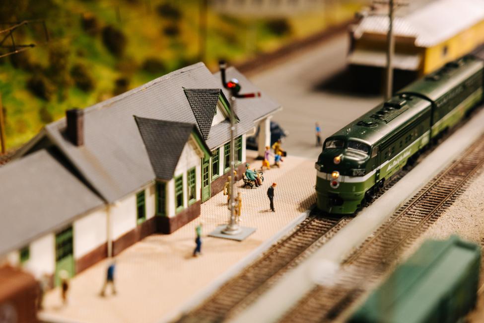 A green model train pulls into a station with tiny people waiting on the platform