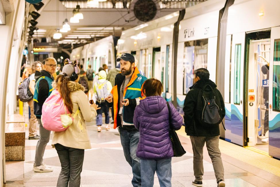 A Sound Transit ambassador in a teal vest talks to passengers at Pioneer Square Station, with a Link train in the background.