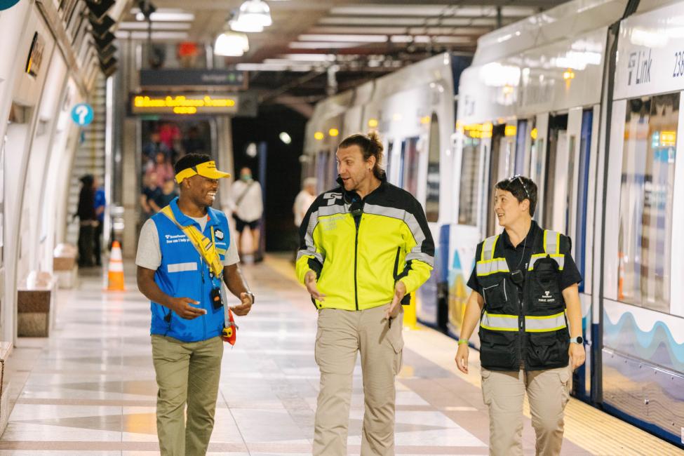 A fare ambassador in a blue vest walks along a Link platform with two security officers