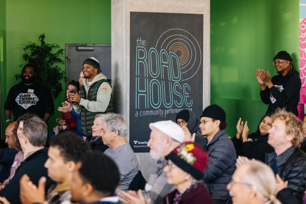 A crowd applauds. A colorful sign that says 'Roadhouse' is in the back of the room, behind the seated people