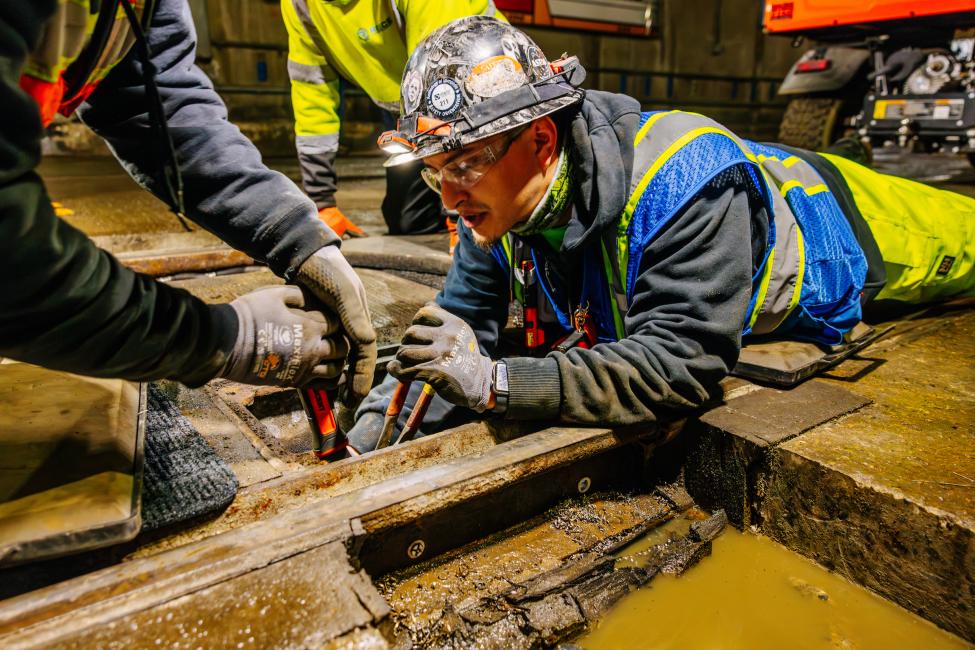 A worker in protective gear lays on his stomach to look at something 