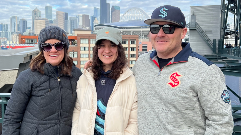 Three people smile with their Kraken gear on and downtown Seattle buildings in the background