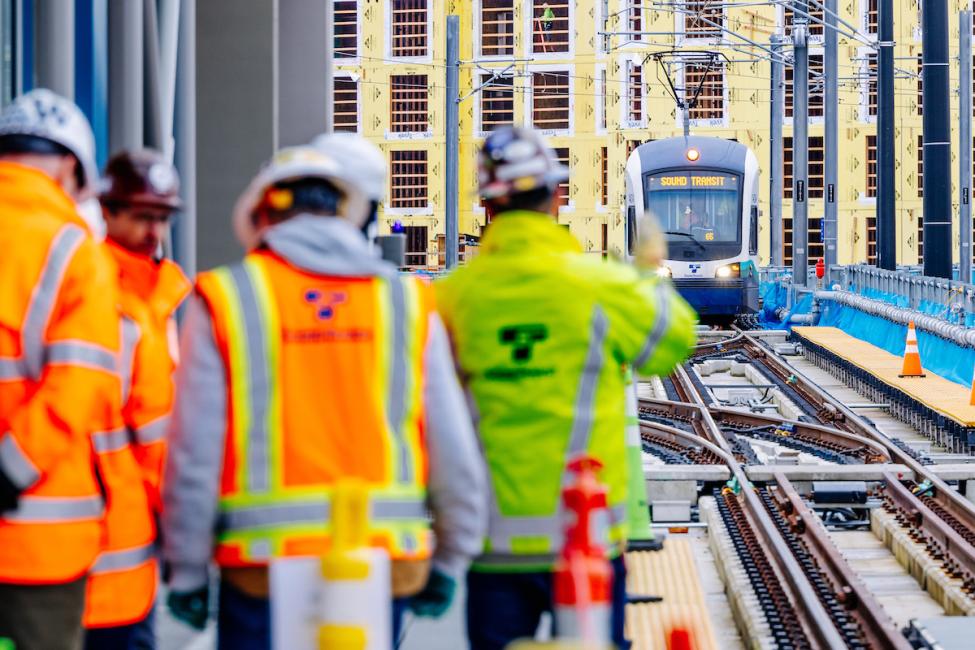 People in Sound Transit reflective jackets and hard hats look down the tracks toward an approaching Link train