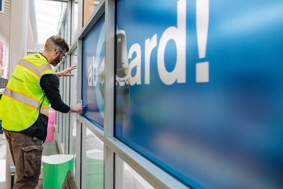 A worker in a yellow vest installs art on a 2 Line station to get ready for the opening celebration