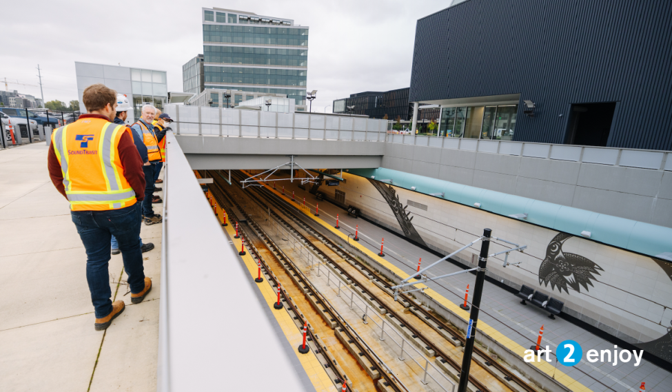 People in orange vests look down at the Spring District Station platform, which features art by Louie Gong
