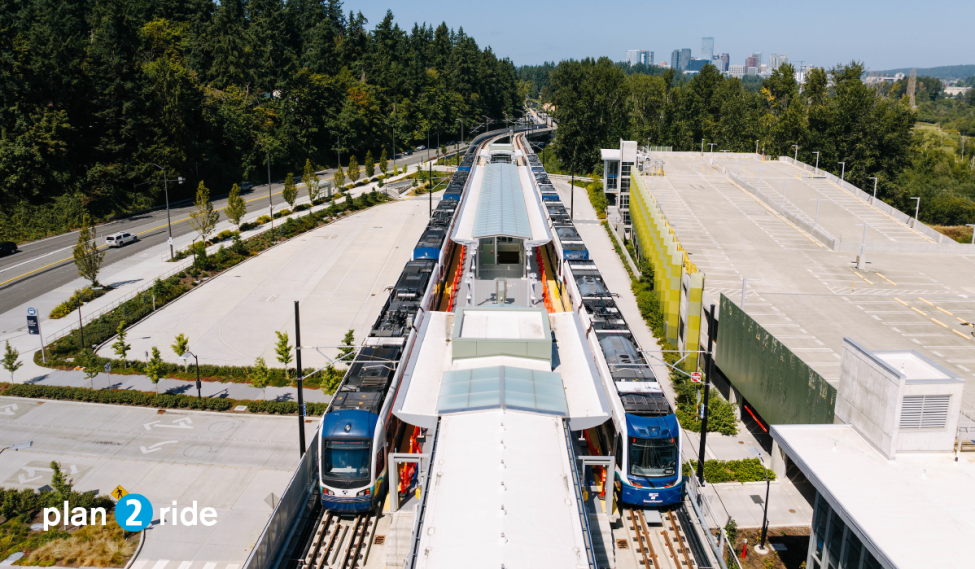 Two trains at South Bellevue Station on a sunny day, with downtown Bellevue in the background. Text reads 'plan 2 ride'