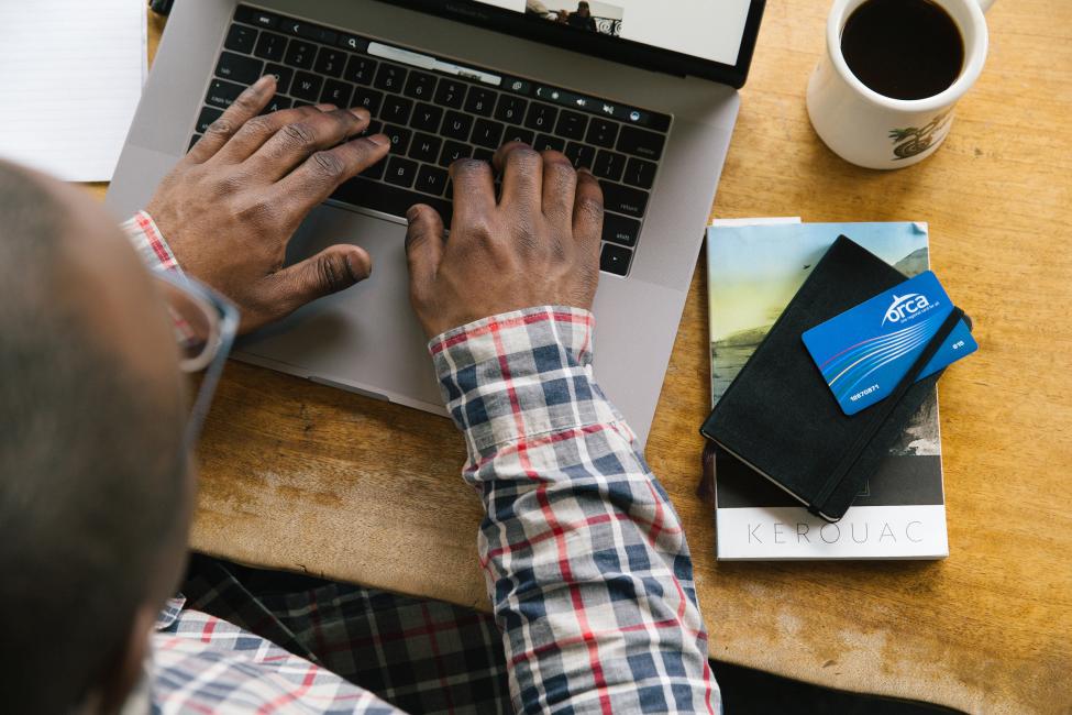 A person types on a laptop with a cup of coffee, notebook and ORCA card nearby on their desk.