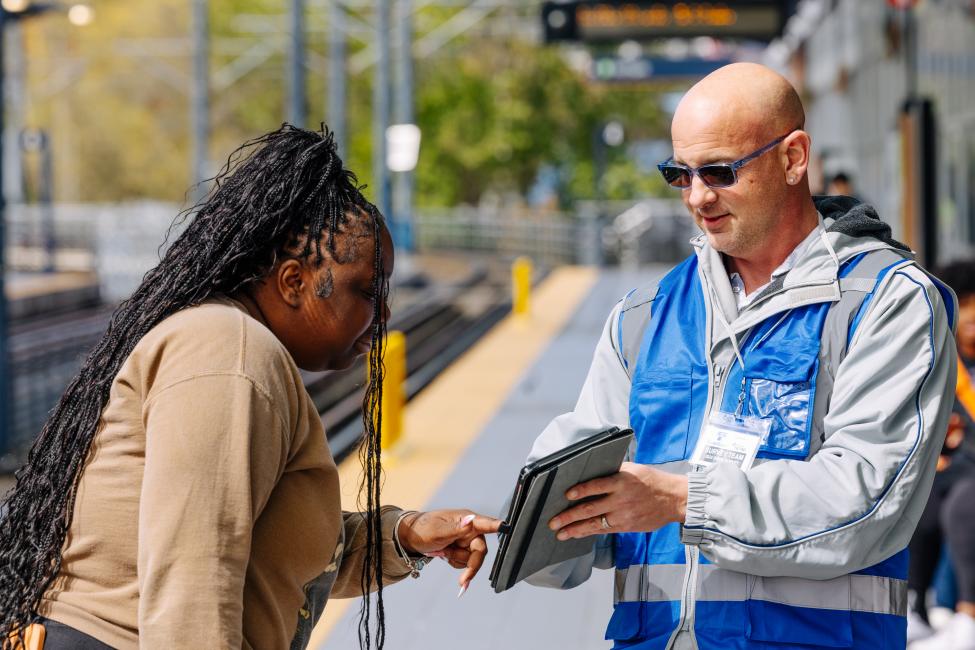 A surveyor wearing a blue vest talks to a Sound Transit passenger on a train platform