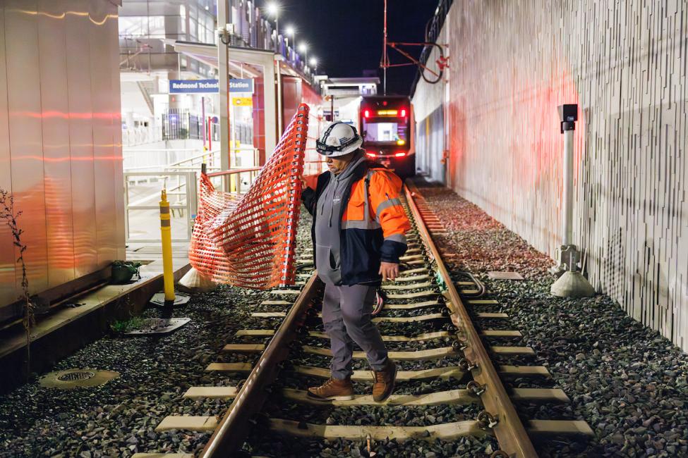 A construction worker in an orange jacket walks along train tracks in front of an orange fence