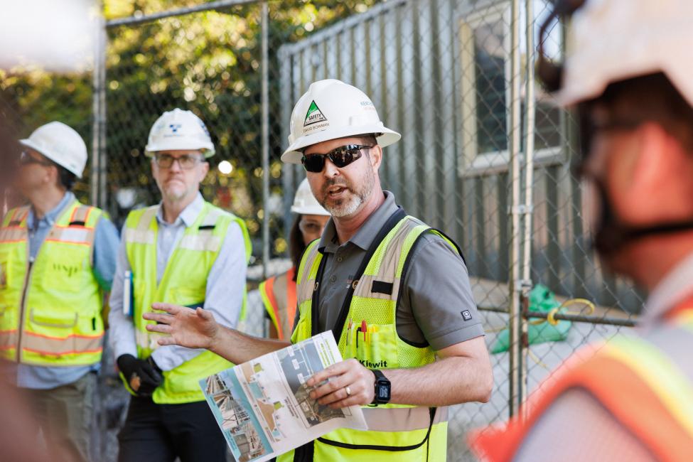 A person wearing a white hard hat and yellow reflective vest talks to a group of people