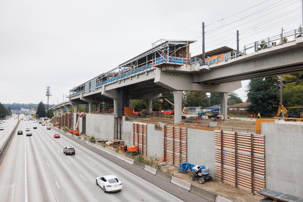 NE 130th St is a construction zone, with the roof of a new light rail station being built while trains pass on the tracks beneath it. The station is right next to I-5 in north Seattle.