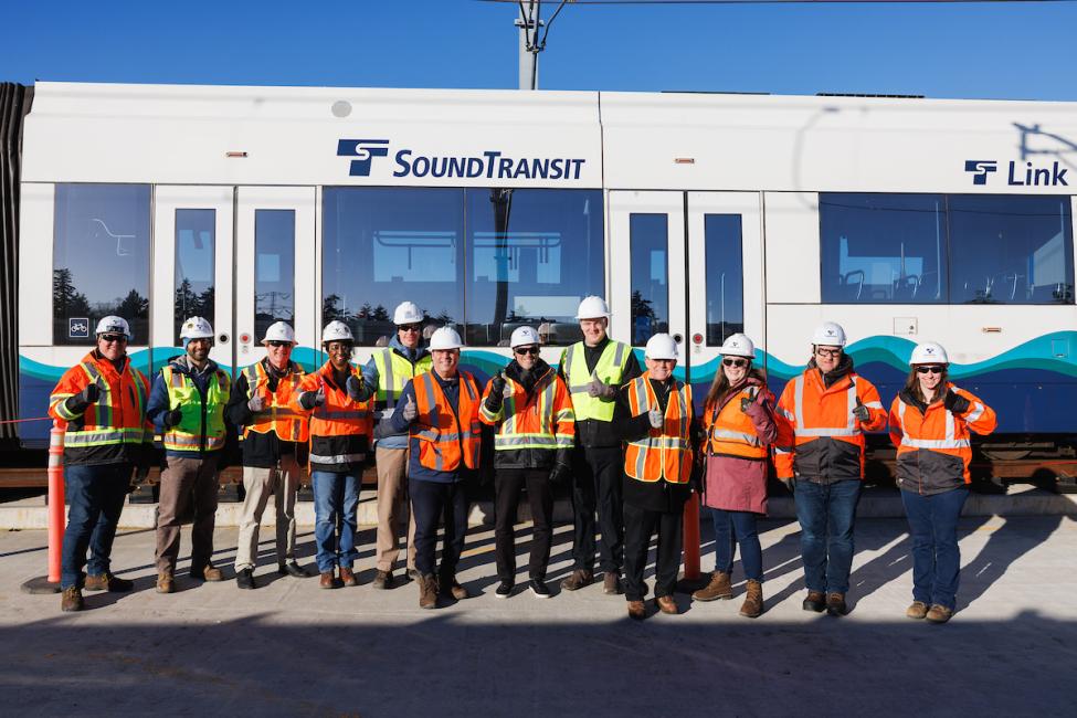 A crew of Sound Transit employees wearing bright orange protective gear smile and wave with a Link train in the background.