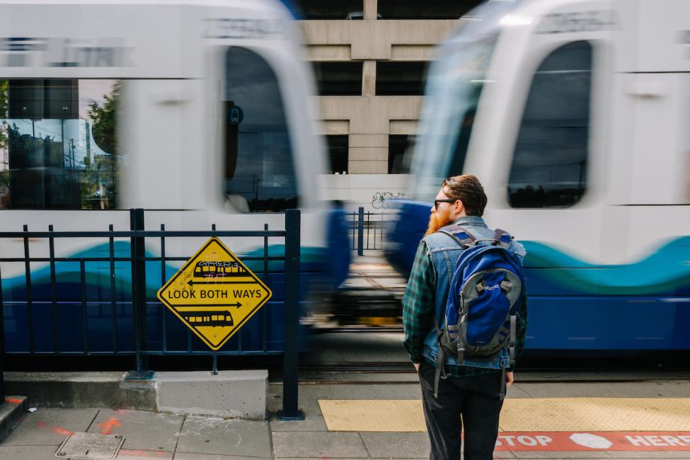 A passenger waits for a Link train to pass before crossing. A yellow sign to his left reads 'look both ways.'