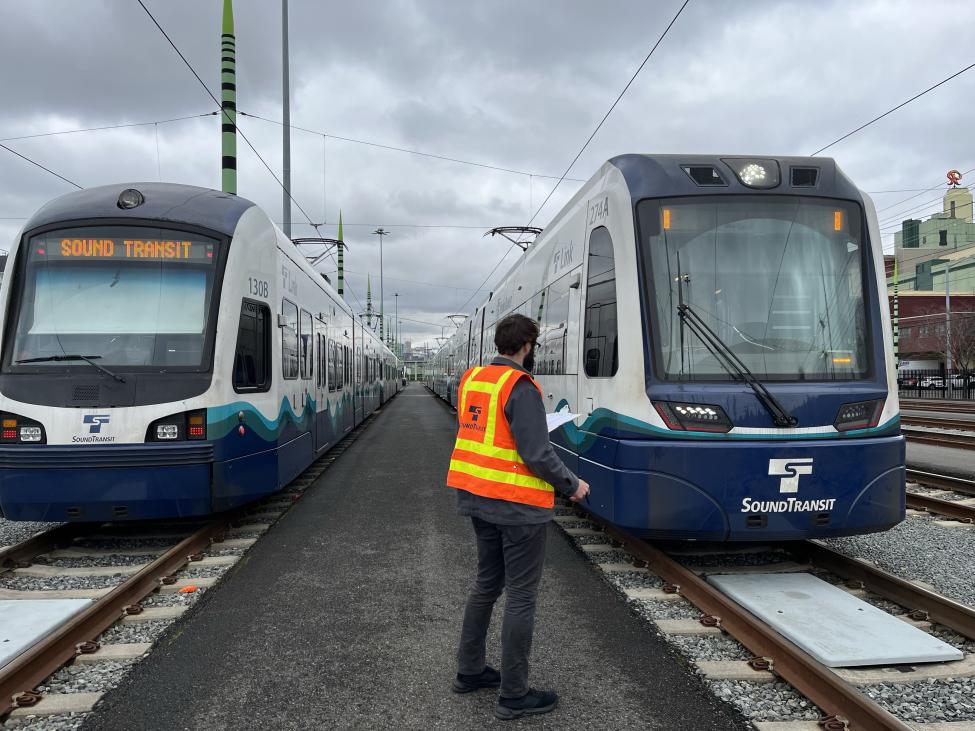 A person wearing an orange vest stands between two Link light rail vehicles