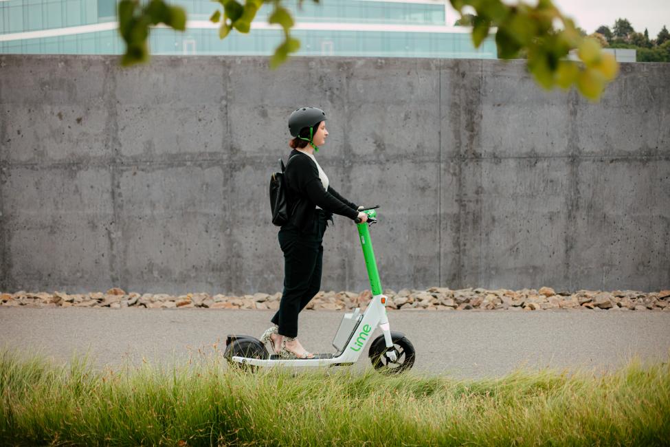 Side angle of a woman riding a lime scooter with a concrete wall behind her.