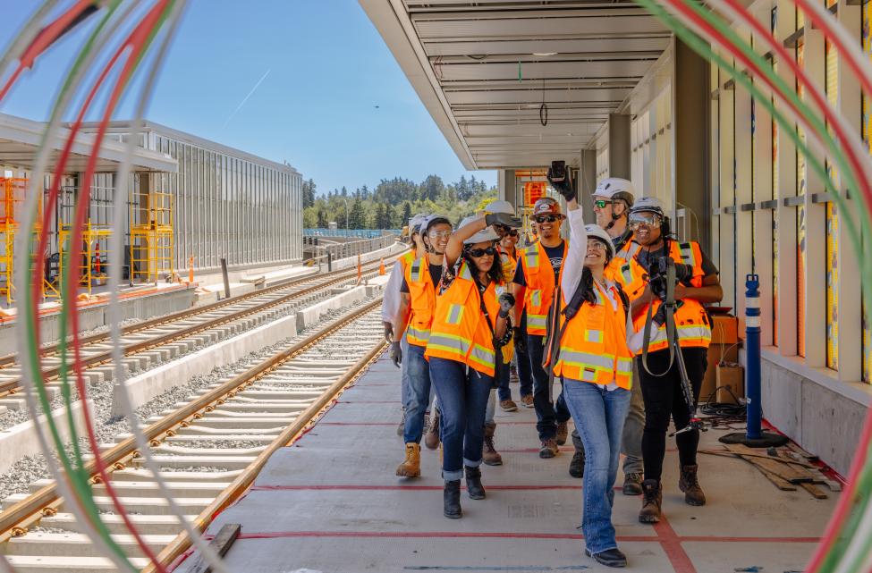 Eight Sound Transit employees wearing hard hats and orange vests pause for a selfie during a tour of a new light rail station