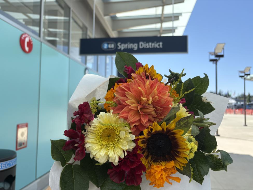 A bouquet of flowers, including pink dahlias and sunflowers, with a sign for the entrance to Spring District Station in the background.