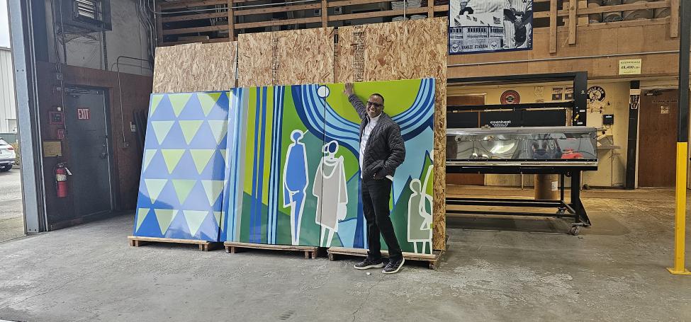 Artist Yeggy Michael stands in front of blue and green panels in a fabrication space