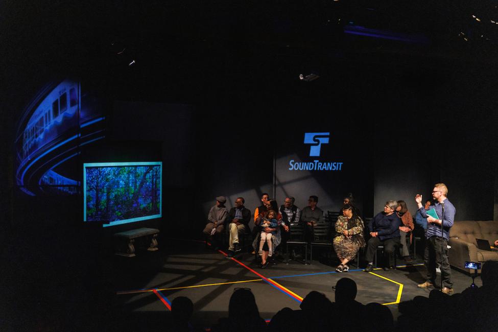 Cable Griffith speaks about his art at Downtown Redmond Station on a darkly lit stage with other artists behind him