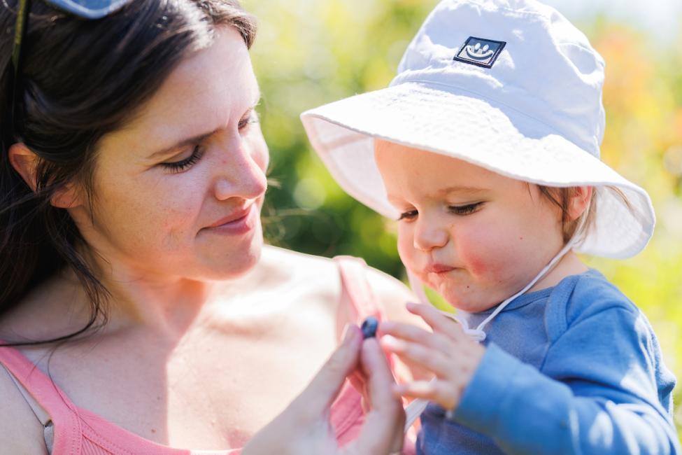 A baby wearing a white hat reaches out for a blueberry held by mom, wearing a pink shirt