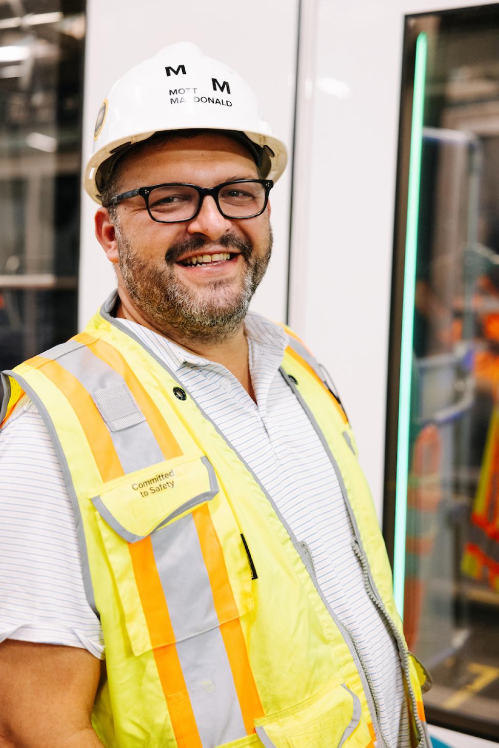 A person wearing a white hard hat, striped shirt and yellow vest smiles with a Link train in the background