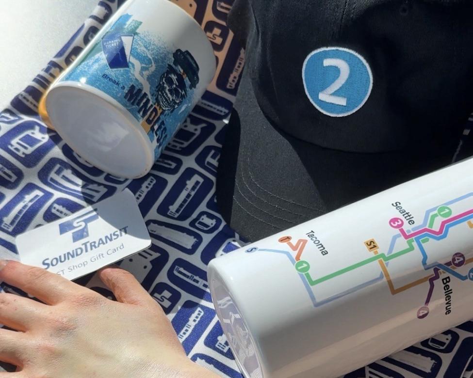 A collection of items from the Sound Transit shop including a mug, tote, 2 Line hat, water bottle and gift card.