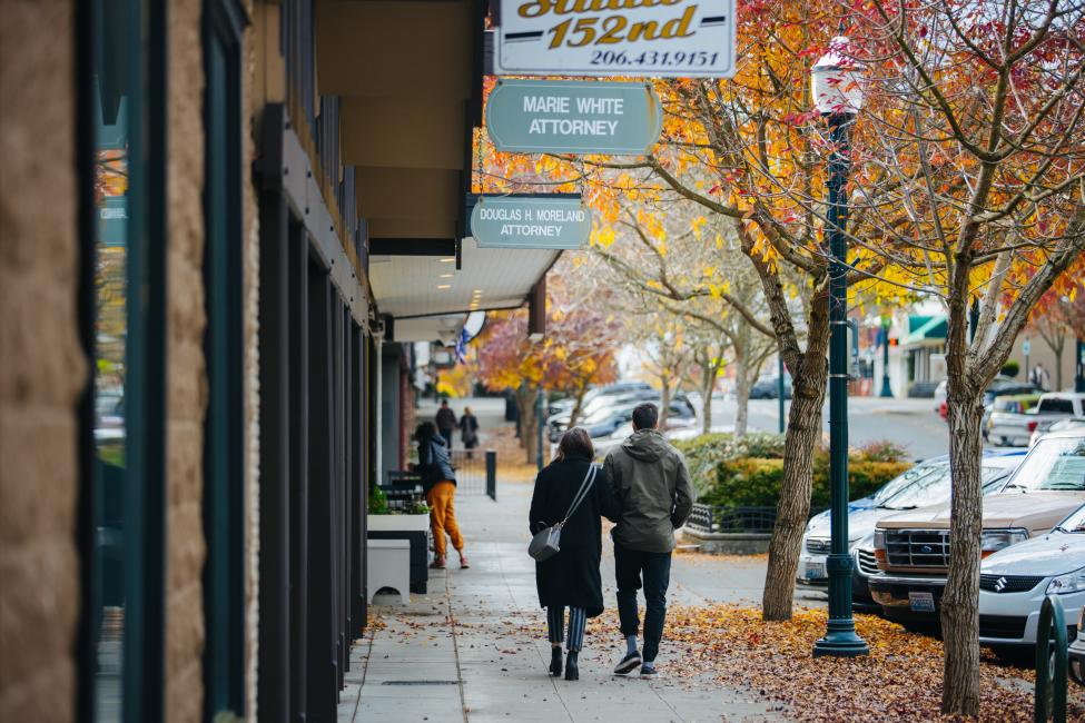 Two people walk on a sidewalk in downtown Burien amidst trees with orange and yellow leaves