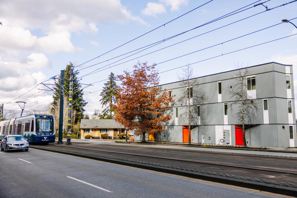 Gray colored housing with red doors on a street with cars and a light rail track. 