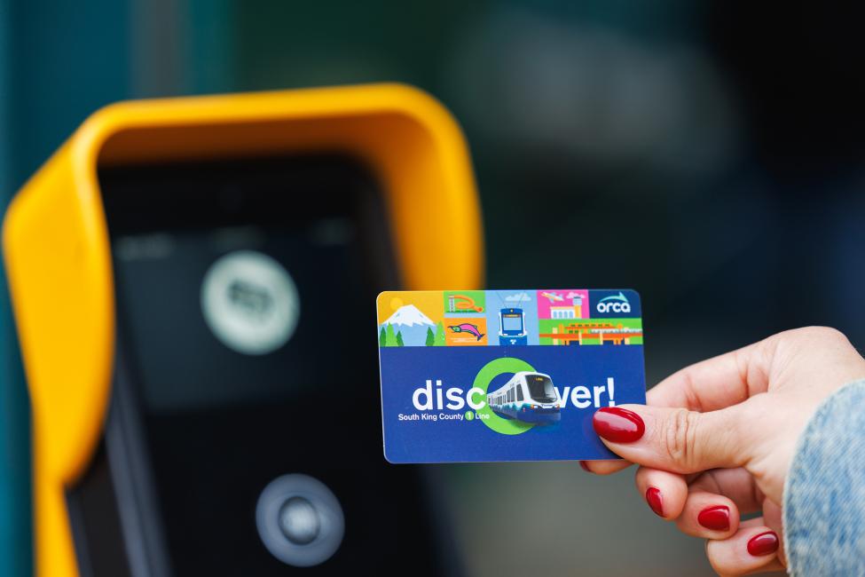 A hand holds an ORCA card up to a yellow tapper. The card says "Discover" and has other designs that commemorate the 1 Line extension to Federal Way
