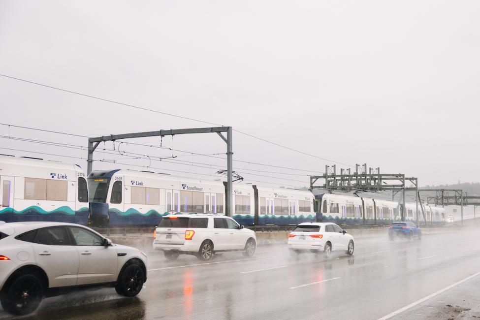 Cars drive on the I-90 floating bridge in the rain, with a Link train also running on the bridge to the left of them 